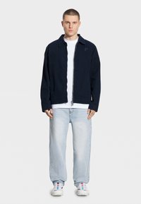 Granatowa kurtka zapinana na zamek z kołnierzykiem, kieszeniami na froncie i haftowanym logo, w zestawieniu z białym t-shirtem, jasnoniebieskimi dżinsami i trampkami.