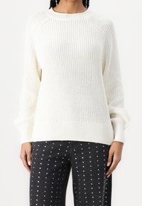 Pull blanc tricoté avec un col et des poignets côtelés, présentant une coupe ample et un motif texturé, associé à un jean noir orné de clous argentés.