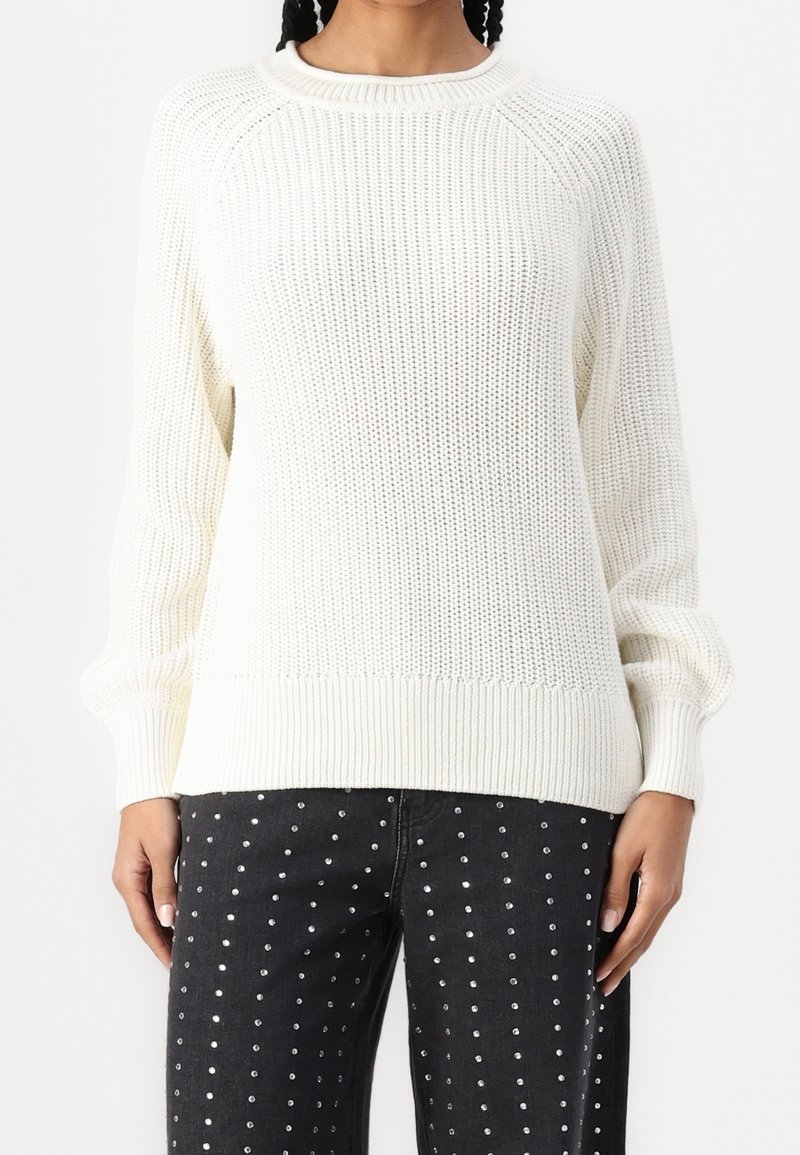 Pull blanc tricoté avec un col et des poignets côtelés, présentant une coupe ample et un motif texturé, associé à un jean noir orné de clous argentés.