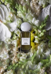 Butelka perfum z prostokątnym szklanym korpusem, białą okrągłą nakrętką, otoczona białymi i zielonymi kwiatami, cechująca się delikatnymi florystycznymi teksturami i kolorami.