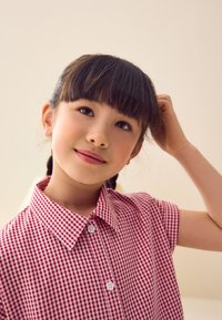 Camicia a maniche corte con collo, fantasia a quadri rossi e bianchi, con chiusura a bottoni e tessuto lavorato. Sfondo liscio e chiaro.