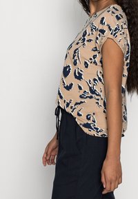 Beige topp med kort ärm och marinblå blommönster, avslappnad passform, rundad fåll och dragsko vid midjan. Slät tygkänsla.