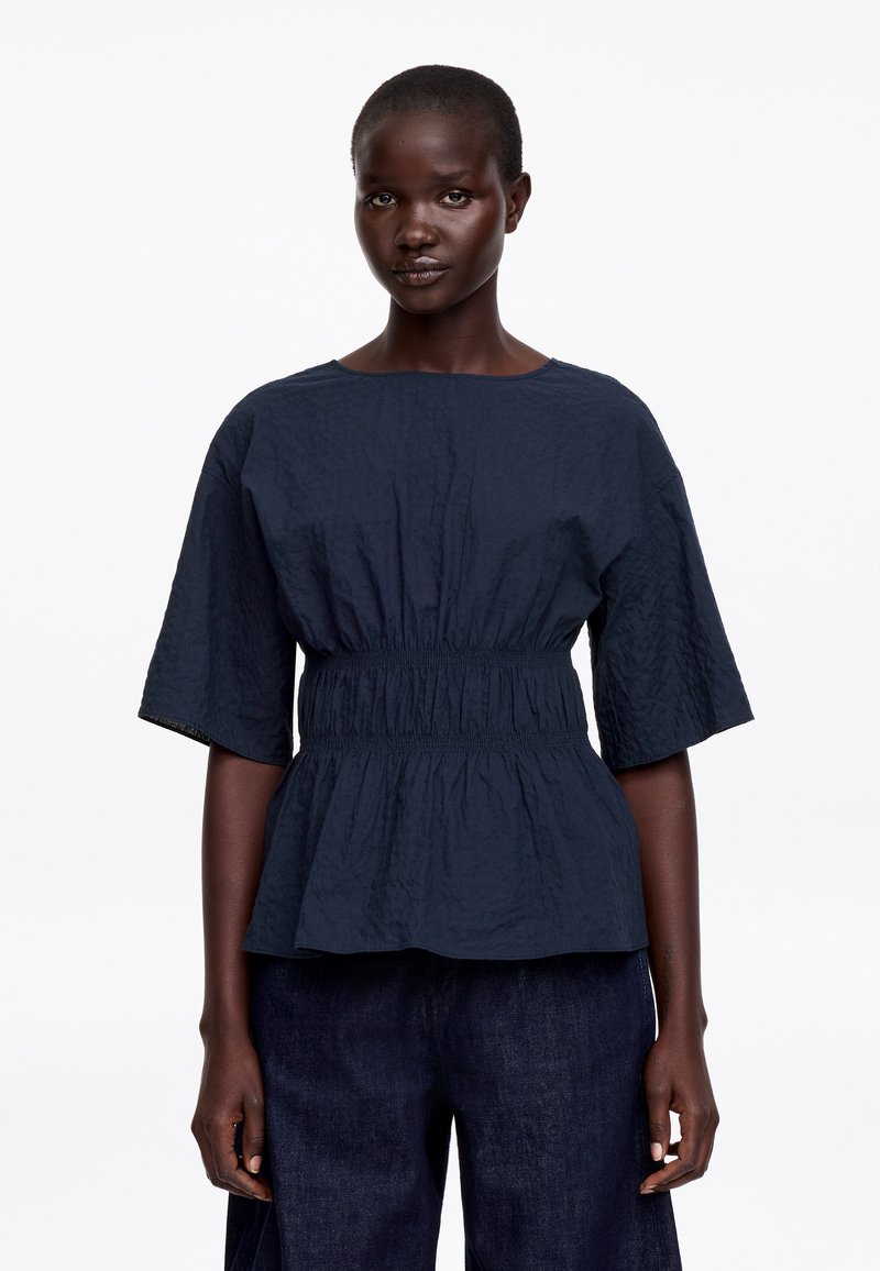 Femme portant un chemisier texturé bleu marine à manches larges et taille froncée, associé à un pantalon large bleu foncé, debout devant un fond blanc.
