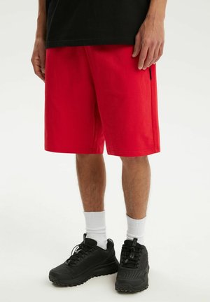 Osoba nosząca jaskrawe czerwone szorty, białe skarpety sportowe, czarne trampki i czarną koszulkę, stojąca na białym tle.