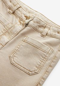 Beige denim kangas, millel on tagatasku detail, nähtavate õmblustega ja väike metallnupp vöökoha lähedal.