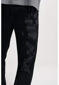 Jeans en denim noir avec des détails déchirés et des bords effilochés. Le design comprend différentes nuances de noir et des zones texturées.