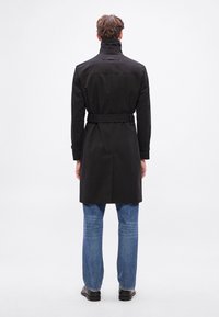 Mustast värvist trentsmantel, millel on kõrge krae, vöökohal vöö ning nööpide detailid. Modell kannab siniseid teksapükse ja musti kingi, vaadatuna tagantpoolt.