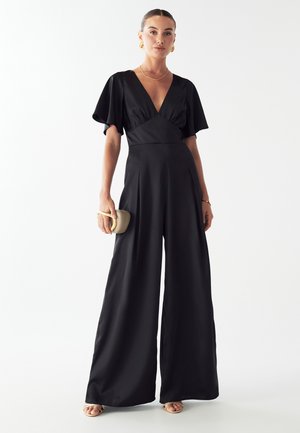 Schwarzer Jumpsuit mit tiefem V-Ausschnitt, kurzen luftigen Ärmeln und weitem Bein. Geschmeidige Textur mit Falten und einer minimalistischen beigen Clutch.