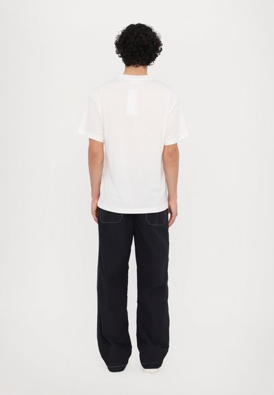 T-shirt en coton blanc à manches courtes, vue de dos. Associé à un jean large noir avec des coutures blanches contrastantes.