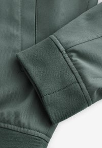 Gros plan sur le poignet d’une veste verte avec un tissu tricoté côtelé et des coutures détaillées sur la manche et la couture de la poche.