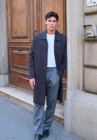 Mand i mørk frakke, hvid skjorte, grå bukser og sorte sko står på fortovet ved en trædør og en stenbygning.