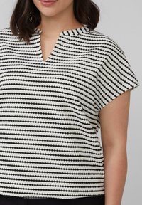 Vrouw in een zwart-wit gestreepte top met korte mouwen en een kleine V-halsuitsparing, weergegeven van hals tot taille.