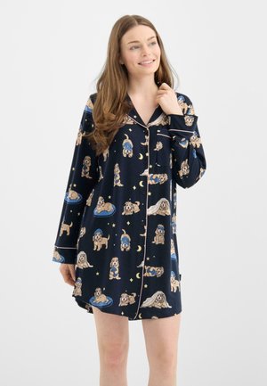 Ung kvinde smilende iført en marineblå natkjole med forskellige legende hundeillustrationer og stjerner, stående foran en ensfarvet baggrund.