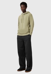 Khaki sweatshirt z kapturem z przednią kieszenią, połączony z luźnymi czarnymi spodniami i czarnymi butami z brązową podeszwą.