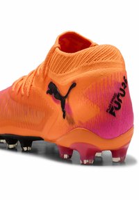 Puman jalkapallokengät kirkkaan oransseina, joissa liukuvärjätyt vaaleanpunaiset yksityiskohdat, teksturoitu synteettinen päällinen, joustava kaulus ja musta brändäys sivulla.