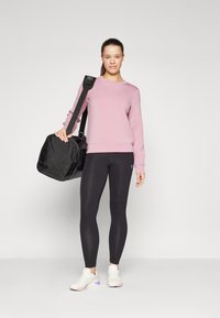 Ružová mikina, čierne legíny a biele tenisky. Model drží čiernu športovú tašku s dlhým popruhom. Elegantný, moderný dizajn a strih.