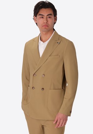 Uomo che indossa una giacca doppiopetto beige con grandi tasche e una camicia bianca, in piedi davanti a uno sfondo bianco.