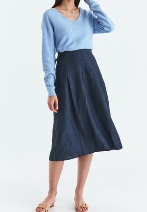 Kvinde iført en lyseblå V-hals sweater, et mørkeblåt A-formet midiskørt og beige vævede slidssandaler, stående med den ene hånd berørende hendes hår.