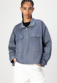 Lett, blågrå zip-up jakke med ståkrage, to frontlommer og avslappet passform. Glatt tekstur; minimale designdetaljer.