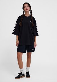 Zwarte oversized t-shirt met witte chevronstrepen op de mouwen, gecombineerd met zwarte shorts. Model draagt zwarte sneakers en witte merksokken.