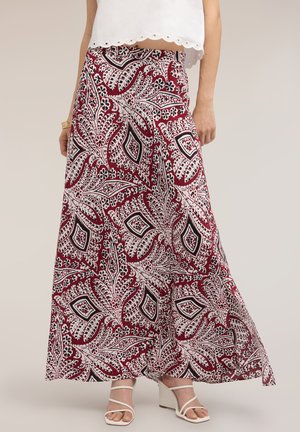 Femme portant une longue jupe bordeaux avec des motifs floraux cachemire blancs et noirs et des sandales compensées blanches à brides.