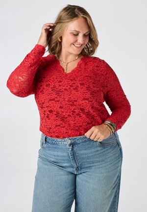 Vrouw glimlacht en kijkt naar beneden terwijl ze haar haar aanraakt, draagt een rood kanten top met lange mouwen en lichtblauwe high-waisted jeans.