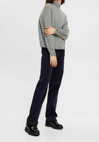 Pull en maille grise avec un col montant et un motif texturé, avec des accents de boutons sur les manches. Associé à un pantalon en velours côtelé bleu marine et des chaussures noires.