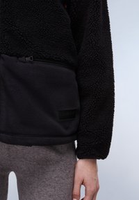Primo piano di una persona che indossa una giacca in pile nera con una tasca con zip e pantaloni grigi, con la mano rilassata al fianco.