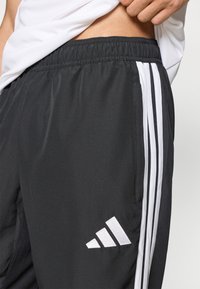 Μαύρο αθλητικό παντελόνι με λευκές κάθετες ρίγες και κεντημένο λογότυπο. Το ύφασμα είναι λείο με ελαφρώς υφή επιφάνειας.