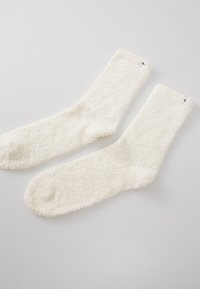 Λευκές, φουσκωτές κάλτσες με απαλή υφή και ριγωτό λάστιχο. Διαθέτουν μια μικρή ετικέτα με λογότυπο στο πλάι. Χωρίς διακριτά σχέδια ή αξεσουάρ.