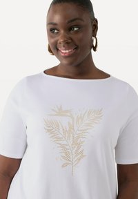 Camiseta blanca de manga corta con diseño de hoja en foil dorado y pequeños acentos de tachuelas, material de algodón y escote redondeado.