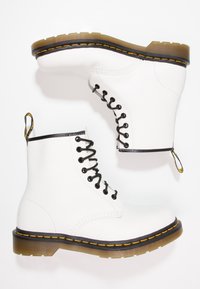 Botas de tobillo de cuero blanco con cordones negros, costuras amarillas y suela de goma. Cuentan con una lengüeta en el talón y un diseño de punta redonda.