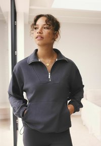 Tmavě šedý pulovr s vysokým límcem, polovičním zipem vpředu a elastickými manžetami. Obsahuje jemné logo a přední kapsu s detaily užších šňůrek.