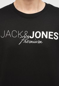 Μαύρο μπλουζάκι με λευκό κείμενο "JACK & JONES" σε έντονα γράμματα και γραφή "premium" που επικαλύπτει το και (&) στο πάνω μέρος του στήθους ενός ατόμου.