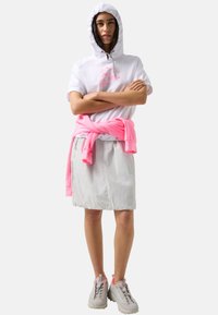 Joven mujer con un top blanco con capucha y mangas cortas, falda gris claro, sudadera rosa atada a la cintura y zapatillas blancas con detalles en rosa.