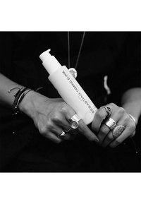 Weiße Plastikpumpe mit dem Etikett "Sebastian Taming Elixir", gehalten zwischen Händen, die mit mehreren silbernen Ringen geschmückt sind. Schwarzer Hintergrund.