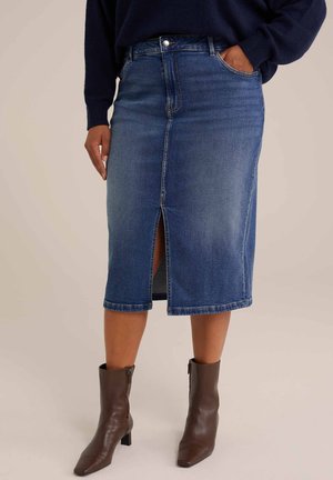 Jupe en denim bleu moyen avec une fente frontale, taille haute et design standard à cinq poches. Associée à des bottines brunes.