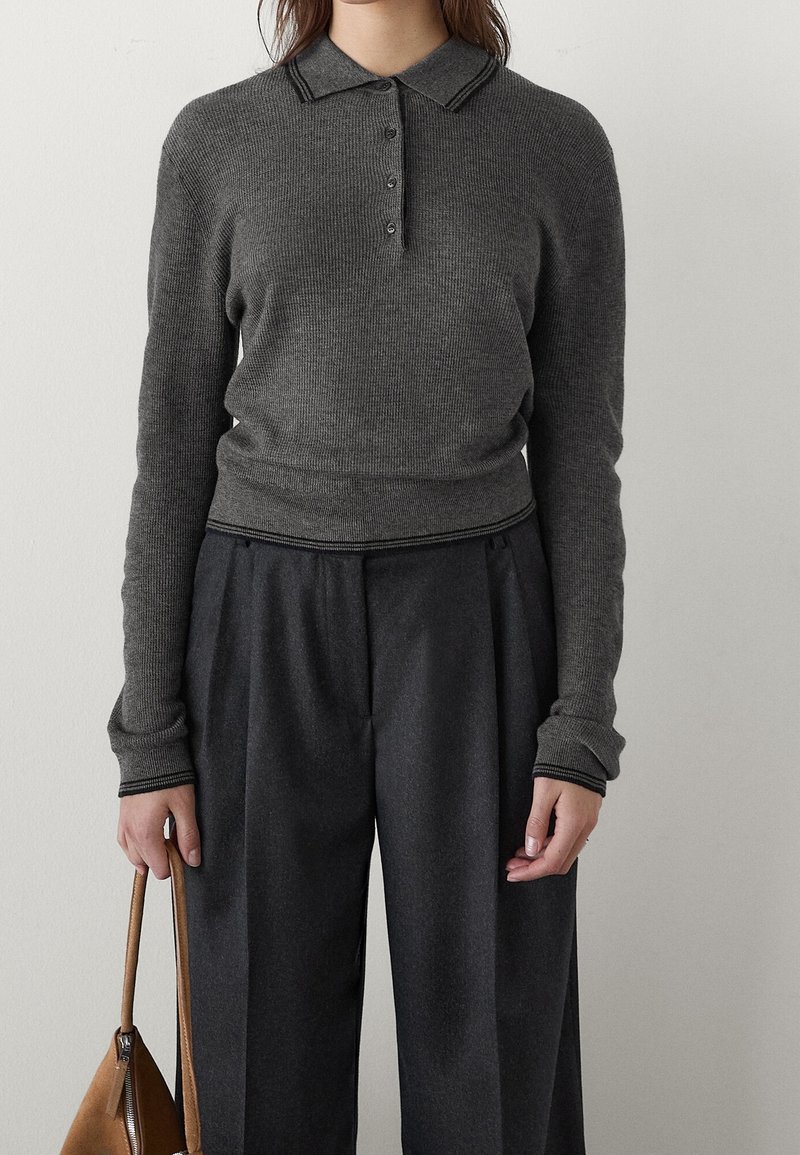 Femme portant un pull polo cintré à manches longues en maille côtelée gris foncé et un pantalon large taille haute couleur charbon, tenant un sac à main marron.