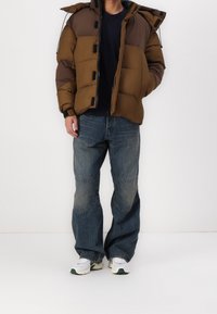 Braune und dunkelbraune Steppjacke mit Kapuze, die große ge quiltete Abschnitte und vordere Druckknöpfe aufweist, kombiniert mit lockeren blauen Jeans und weißen Turnschuhen.