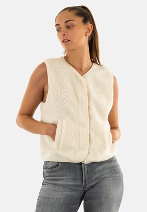 Young woman with ponytail wearing a sleeveless cream fleece vest and gray jeans, hands in vest pockets, looking to the side.