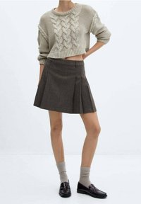 Przycięty beżowy sweter w warkocze, w zestawieniu z brązową spódnicą plisowaną. Modelka ma na sobie jasnoszare skarpetki i ciemne mokasyny. Prosty, teksturowany design.