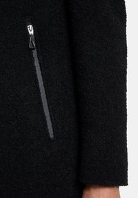 Cappotto nero texturizzato con una tasca con zip e hardware nero lucido sul lato, con una finitura esterna morbida e pelosa.