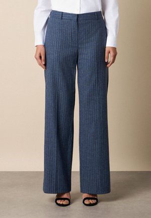 Personne portant un pantalon large à fines rayures bleues, une chemise blanche et des sandales noires à bout ouvert, debout devant un fond beige.