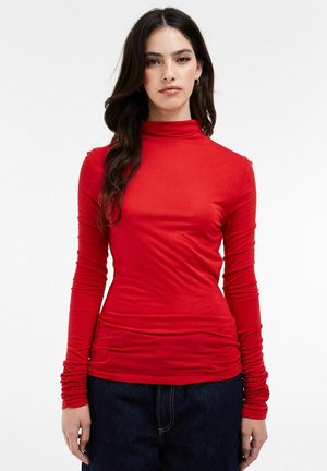 Femme aux longs cheveux foncés portant un pull rouge ajusté à col roulé avec des manches froncées et un jean bleu foncé, debout devant un fond blanc uni.