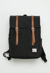 Sac à dos en toile noire avec des sangles en cuir marron et des boucles en métal. Comprend un écusson logo, deux poches latérales et des bretelles réglables.