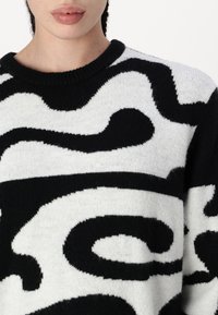 Pull en tricot noir et blanc avec un motif ondulé, col rond et finition texturée. Accent sur les couleurs contrastées et les détails de design.