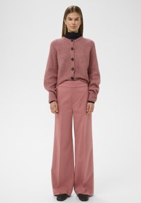 Egy nő poros rózsaszín gombos kardigánt visel fekete garbón, széles lábú poros rózsaszín nadrágban és fekete cipőben, egy sima háttér előtt áll.