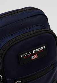Borsa in nylon blu navy con cerniere nere e un'etichetta rettangolare con scritto "POLO SPORT." Presenta una superficie texture e una forma rettangolare piatta.