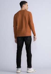 Maglione di un colore ruggine, con texture, maniche lunghe, abbinato a pantaloni neri e scarpe da ginnastica bianche, visto da dietro su uno sfondo chiaro.