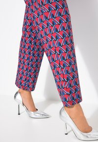 Pantalones coloridos con patrones en rojo, azul y negro con formas abstractas, combinados con unos zapatos de tacón puntiagudo y brillantes en plata.